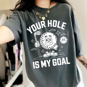 May include: Dark grey t-shirt featuring the text "YOUR HOLE IS MY GOAL" and a cartoon golf ball character holding a golf club. The design includes clouds, stars, and the words "RIM SERVICE".