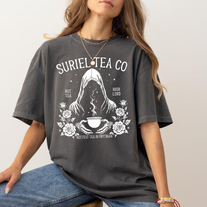 May include: Dark gray t-shirt with a white graphic featuring the text "SURIEL TEA CO" above an illustration of a hooded figure holding a teacup. The design includes roses and the phrases "HOT TEA", "HIGH LORD", and "HOTTEST TEA IN PRYTHIAN".