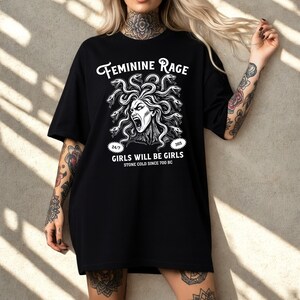 May include: Black t-shirt featuring a white graphic of Medusa with snakes for hair, screaming. The text "FEMININE RAGE" is above the graphic, with "GIRLS WILL BE GIRLS" and "STONE COLD SINCE 700 BC" below. The shirt is a casual, oversized style.