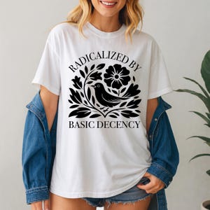 May include: White t-shirt with the text "RADICALIZED BY BASIC DECENCY" in a black, stylized font. The design includes a black bird, flowers, and leaves. The shirt is a casual, everyday wear item.