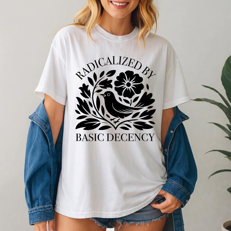May include: White t-shirt with the text "RADICALIZED BY BASIC DECENCY" in a black, stylized font. The design includes a black bird, flowers, and leaves. The shirt is a casual, everyday wear item.