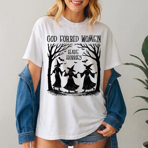 K&ouml;nnte beinhalten: Wei&szlig;es T-Shirt mit dem schwarzen Text "GOD FORBID WOMEN HAVE HOBBIES". Die Grafik zeigt drei Silhouetten von Hexen, die sich an den H&auml;nden halten, mit Flederm&auml;usen und B&auml;umen.