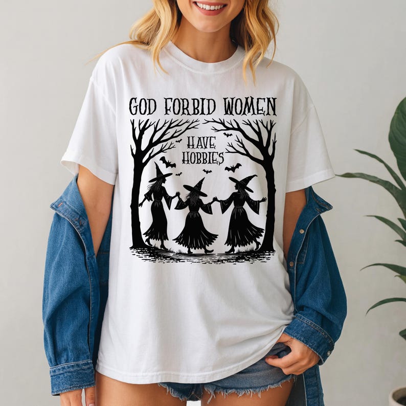 K&ouml;nnte beinhalten: Wei&szlig;es T-Shirt mit dem schwarzen Text "GOD FORBID WOMEN HAVE HOBBIES". Die Grafik zeigt drei Silhouetten von Hexen, die sich an den H&auml;nden halten, mit Flederm&auml;usen und B&auml;umen.