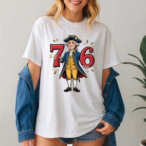May include: White t-shirt with a cartoon illustration of a man in colonial attire holding the numbers 7 and 6. The man wears a blue coat, yellow vest, and tricorn hat. The numbers are red with yellow accents.