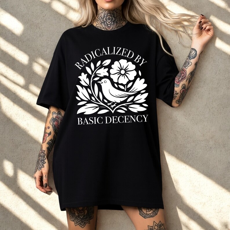 May include: Black t-shirt with a white graphic featuring a bird, flowers, and leaves. The text "RADICALIZED BY BASIC DECENCY" is arched above the design. The shirt has short sleeves and a relaxed fit.