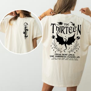 May include: A cream-colored t-shirt with a black graphic print of a dragon and the text "The Thirteen" and "From now until the darkness claims us". The text lists the names of thirteen characters: Manon, Asterin, Sorrel, Vesta, Faline, Fallon, Edda, Briar, Thea, Kaya, Linnea, Ghislaine, and Imogen.