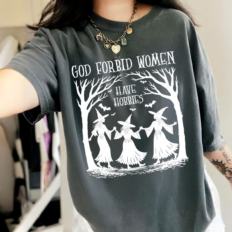 K&ouml;nnte beinhalten: Dunkelgraues T-Shirt mit dem wei&szlig;en Text "GOD FORBID WOMEN HAVE HOBBIES". Die Grafik zeigt drei Hexen, die sich unter stilisierten B&auml;umen an den H&auml;nden halten, mit Flederm&auml;usen.