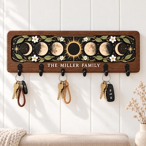 May include: A rectangular wooden wall-mounted key holder featuring a dark brown frame and a central panel displaying a lunar cycle graphic with white flowers and green leaves. The text THE MILLER FAMILY is printed in white below the moon phases.