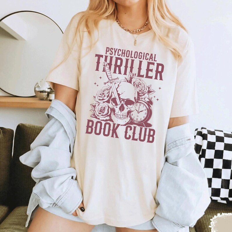 May include: Cream-colored t-shirt with the text "PSYCHOLOGICAL THRILLER BOOK CLUB" in burgundy. The graphic includes a skull, roses, a sword, and a clock. The shirt has short sleeves.