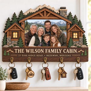 May include: A wooden wall-mounted key holder shaped like a cabin with a central photo frame containing a family portrait. The sign features the text THE WILSON FAMILY CABIN, 27 PINE RIDGE ROAD, BOZEMAN, MONTANA, and includes five brass hooks with keys.