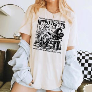 May include: Cream-colored t-shirt with a black graphic design. The design includes text that reads "PROUDLY ANTI-SOCIAL MEMBER OF THE INTROVERTED Book club" with illustrations of a dragon, books, a witch hat, and flowers. The shirt also has the text "MAKING FRIENDS WITH CHARACTERS, NOT PEOPLE."