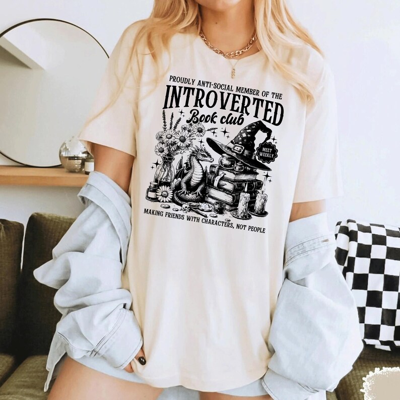 May include: Cream-colored t-shirt with a black graphic design. The design includes text that reads "PROUDLY ANTI-SOCIAL MEMBER OF THE INTROVERTED Book club" with illustrations of a dragon, books, a witch hat, and flowers. The shirt also has the text "MAKING FRIENDS WITH CHARACTERS, NOT PEOPLE."