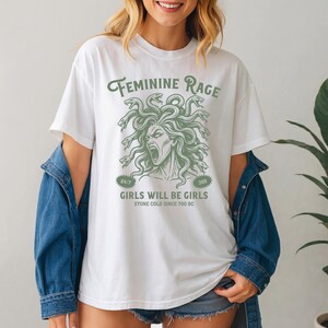 May include: White t-shirt with a green graphic of Medusa's head and the text "FEMININE RAGE." Below the graphic, the text reads "GIRLS WILL BE GIRLS" and "STONE COLD SINCE 700 BC." The shirt has short sleeves and a crew neck.