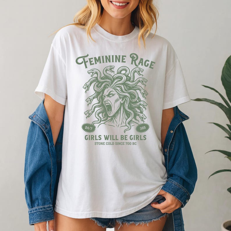 May include: White t-shirt with a green graphic of Medusa's head and the text "FEMININE RAGE." Below the graphic, the text reads "GIRLS WILL BE GIRLS" and "STONE COLD SINCE 700 BC." The shirt has short sleeves and a crew neck.