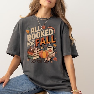 May include: Dark gray t-shirt with the text "ALL BOOKED FOR FALL" and fall-themed graphics. The design includes a stack of books, a pumpkin, a coffee mug, and autumn leaves. The shirt is a comfortable, oversized fit.