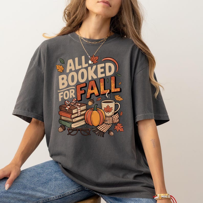 May include: Dark gray t-shirt with the text "ALL BOOKED FOR FALL" and fall-themed graphics. The design includes a stack of books, a pumpkin, a coffee mug, and autumn leaves. The shirt is a comfortable, oversized fit.