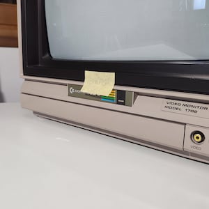 Può includere: Un monitor video Commodore 1702 beige con schermo nero. Il monitor ha un pulsante di accensione e porte di ingresso video/audio. Un post-it giallo è attaccato sul davanti.