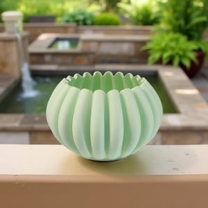 Puede incluir: Un cuenco de cerámica verde claro, acanalado, con un borde festoneado. El cuenco redondo está colocado sobre una superficie beige, con un fondo exterior borroso que incluye una fuente y vegetación.