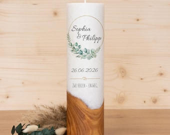 Könnte beinhalten: Eine weiße Säulenkerze mit goldenem Ringdesign, mit den Namen "Sophia & Phillip" und dem Datum "26.06.2026". Die untere Hälfte hat ein Holzmaserungsmuster. Der Text "ZWEI HERZEN - EIN WEG" ist ebenfalls vorhanden.