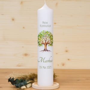 Könnte beinhalten: Eine weiße Stumpenkerze mit einem Baumdesign und dem Text "Meine Kommunion" und "Markus 29. Mai 2025". Die Kerze steht auf einem Holzsockel. Der Baum hat grüne Blätter und einen braunen Stamm.