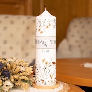 Könnte beinhalten: Eine weiße Säulenkerze mit floralen Mustern und den Namen "Melissa & Leander" sowie dem Datum "17.06.2025". Die Kerze steht auf einem Holzsockel und ist von Trockenblumen umgeben.