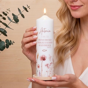 Könnte beinhalten: Eine brennende weiße Kerze mit dem Namen "Antonia" und floralen Illustrationen. Die Kerze trägt den Text "19. APRIL 2026" und einen religiösen Vers. Sie steht auf einem kleinen, runden Holzsockel.