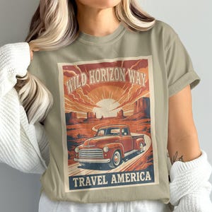 Puede incluir: Camiseta verde oliva con un diseño de póster de viaje vintage. El póster representa una camioneta roja en una carretera desértica, con el texto "Wild Horizon Way" y "Travel America". El diseño tiene una estética retro con temática de viajes.