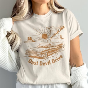 Puede incluir: Camiseta color crema con un gráfico marrón de un descapotable vintage conduciendo por un paisaje desértico con un remolino de polvo. El texto "Dust Devil Drive" está impreso debajo del coche.