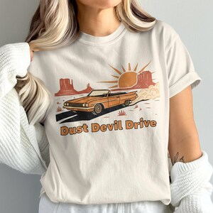 Puede incluir: Camiseta color crema con una escena desértica. Un coche descapotable naranja circula por una carretera. El diseño incluye un sol, montañas y el texto "Dust Devil Drive" en naranja.