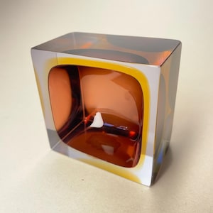 Puede incluir: Un jarrón de vidrio cuadrado con un diseño transparente, blanco y ámbar. El jarrón tiene una abertura rectangular redondeada.