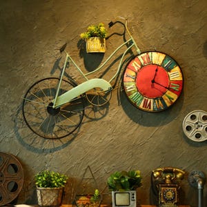 May include: A wall-mounted metal bicycle sculpture featuring a light green frame, a small planter with green foliage, and a large circular clock as the front wheel. The clock face has a red centre, Roman numerals, and a distressed, multi-coloured border.