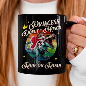 以下が含まれることがあります： 黒いセラミックマグカップ。王冠とマントをつけた猫が恐竜に乗っており、「PRINCESS DONUT & MONGO RIDE OR ROAR」の文字が書かれています。デザインには、虹色の夕日の背景と金色の星が含まれています。
