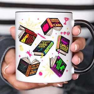 以下が含まれることがあります： 黒いハンドルと縁が付いた白いセラミックマグ。 「Dungeon Crawler Carl」や「The Gate of the Feral Gods」などのタイトルが付いた開いた本のカラフルなイラストが特徴です。 デザインには、ピンクの猫、クモの巣、星が含まれています。