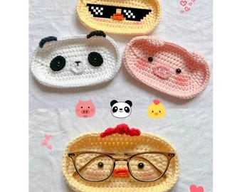 May include: Four crocheted eyeglass holders shaped like animal faces. The holders are yellow, white, pink, and yellow with a pair of glasses on top. Each holder has a different animal face: a chicken, a panda, a pig, and a chicken.
