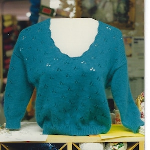 Könnte beinhalten: Ein türkisfarbener Strickpullover mit V-Ausschnitt und einem Lochmuster. Der Pullover ist auf einem Holzregal mit Wollknäueln ausgestellt.