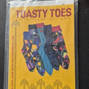 May include: A clear plastic package containing a pattern instruction sheet titled TOASTY TOES. The yellow sheet features a grid background, text, and an illustration of five socks with various colorful geometric and abstract patterns.