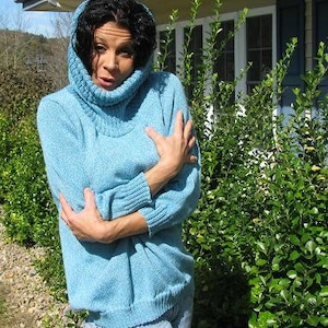 May include: A woman wearing a light blue turtleneck sweater with a folded collar. The sweater is made of a soft, knitted fabric. The woman is standing in front of a green bush.
