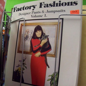 May include: A book titled "Factory Fashions: Designer Pants & Jumpsuits, Volume 1." The cover features a woman in a red dress holding a black cat. The book is on sale.