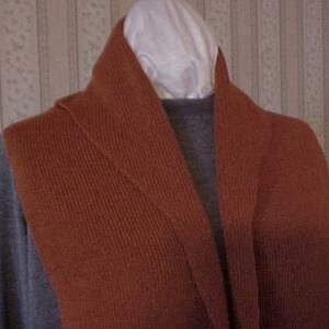 May include: A rust-colored knitted scarf with a long, flowing design. The scarf is folded in half and draped over a gray shirt.