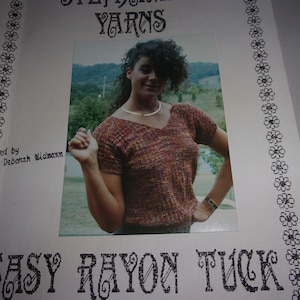 Può includere: Una pagina stampata con una foto di una donna che indossa un top in maglia a maniche corte con scollo a V. Il top è un mix di filati marroni, arancioni e gialli. Il testo "EASY RAYON TUCK" è stampato in basso. Il testo "Modeled by Deborah Widmann" è stampato sul lato sinistro.