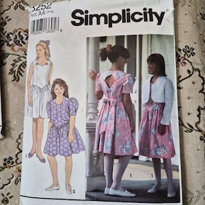 May include: A Simplicity sewing pattern envelope 3252 size AA (7-10) featuring illustrations of dresses with puff sleeves, a back cutout, and a cropped jacket. The cover displays four variations of the garment in floral and solid white fabrics.