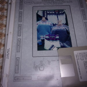 May include: A close-up of a floppy disk with the text "Gini Woodward Publications 1992" and a list of knitting patterns. The disk is labeled "Track 1" and "Track 2" with different knitting patterns listed for each track. The disk is on a white background.