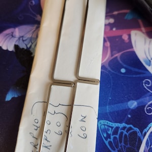 May include: Two silver metal needle threaders with a hook at the end. The threaders are laying on a white surface with handwritten text that says "SRP 40 SKP 50 60 60M".