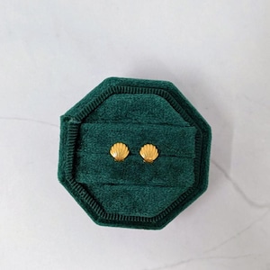 May include: A pair of gold-colored seashell stud earrings displayed in a dark green velvet octagonal jewelry box. The earrings are small and detailed, with a textured surface. The box is open, revealing the earrings.
