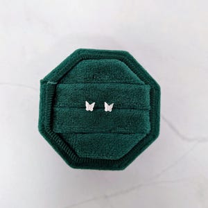 May include: A pair of small, silver butterfly earrings displayed on a dark green velvet jewelry box. The earrings are positioned in the center of the box, showcasing their delicate design. The box is octagonal and has a soft, textured appearance.