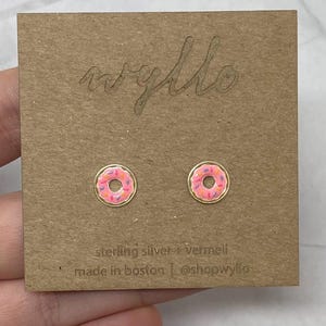 Puede incluir: Un par de pendientes de donut dorados con glaseado rosa y coloridos espolvoreados. Los pendientes están en una tarjeta marrón con el texto "wyllo" y "sterling silver + vermeil" y "made in boston | @shopwyllo".