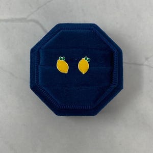 May include: A pair of lemon-shaped earrings in a blue velvet jewelry box. The earrings are yellow with green leaves.