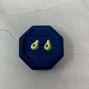 May include: A pair of avocado-shaped earrings in a blue velvet jewelry box. The earrings are green and yellow with a brown seed.