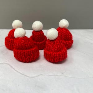 May include: Five red knitted hats with white pom-poms. The hats are small and would be suitable for decorating a Christmas tree or other holiday crafts.