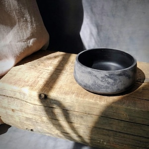 May include: A small, round, dark gray bowl with a textured surface. The bowl sits on a weathered wooden plank, with a neutral-colored fabric in the background. The bowl's interior is a solid black color.